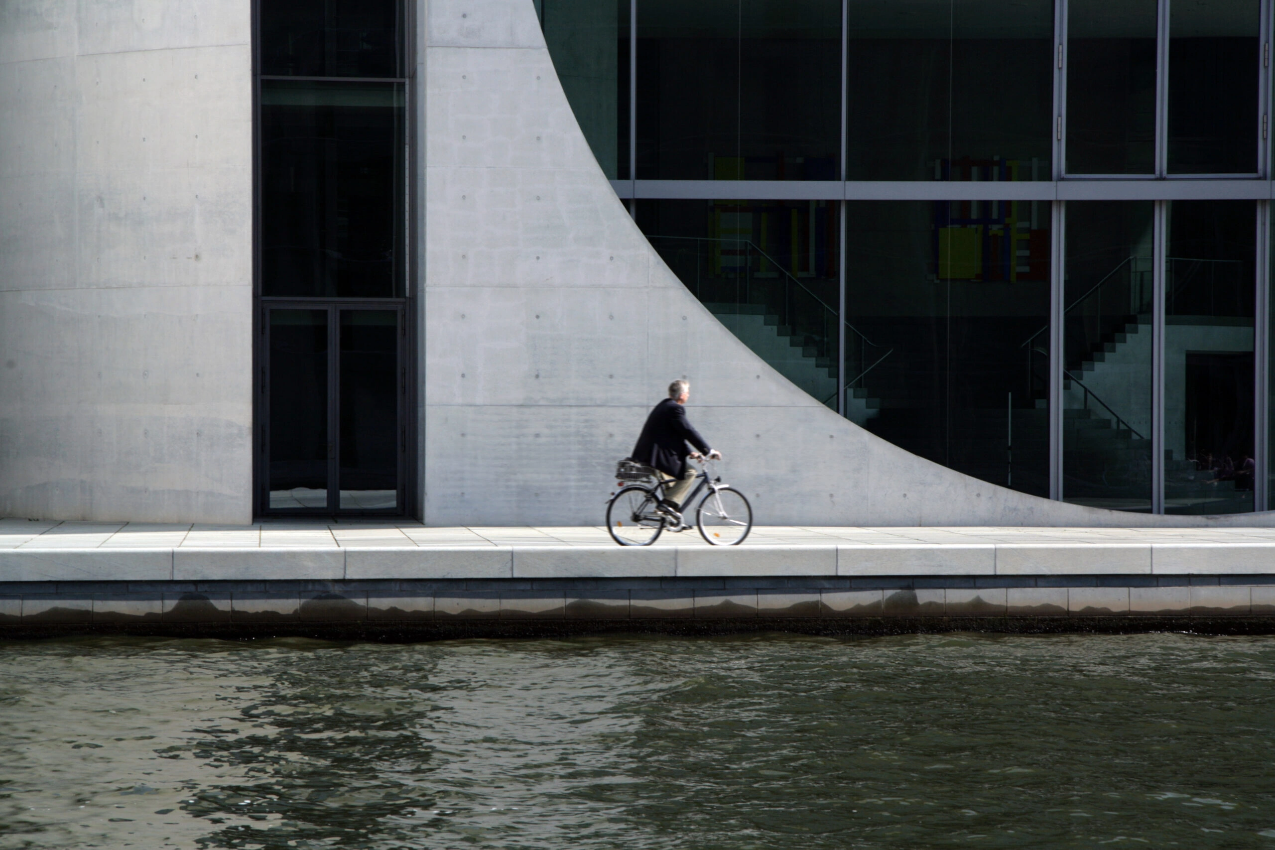 Artistic shot of a businessman on a bicycle with curved architectural wall in the background.