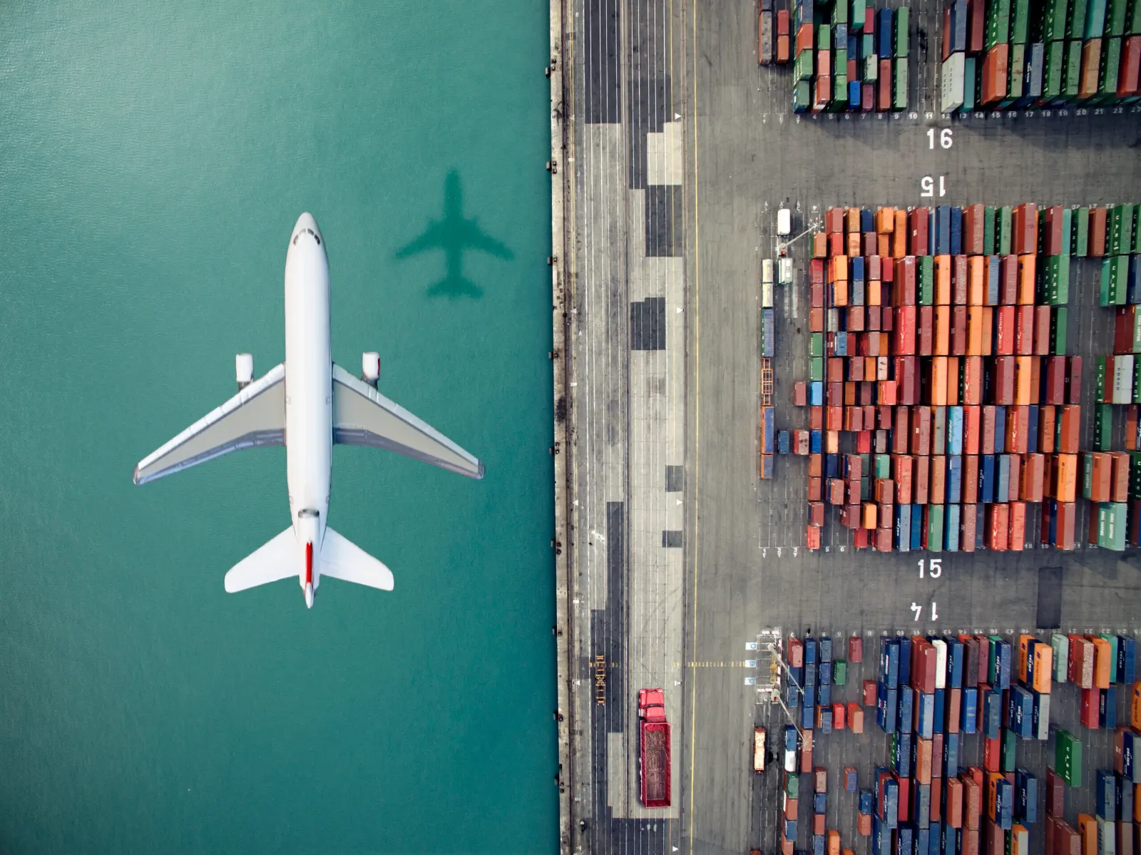 An overhead view of a jet plane flying over a waterway with colorful shipping containers stacked nearby.
