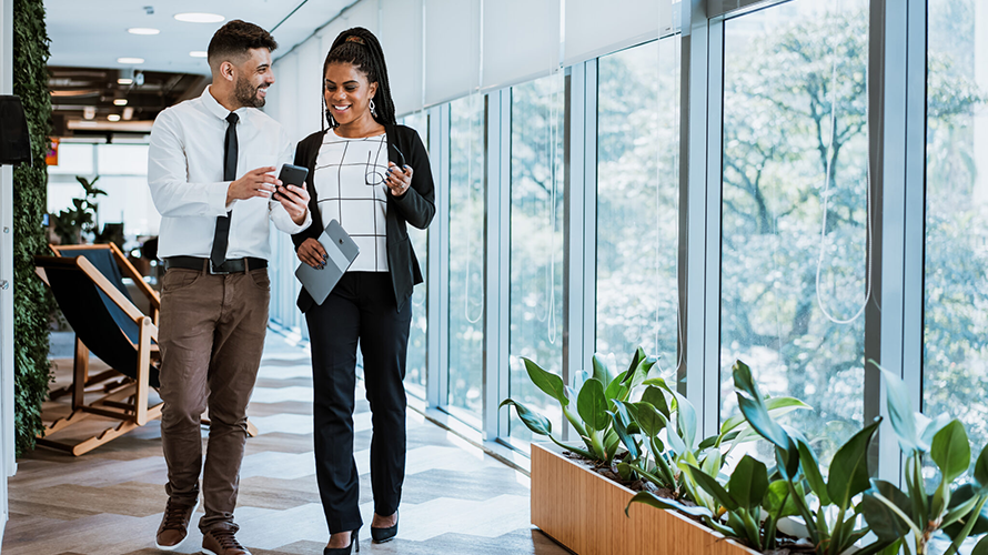 Young businessman and businesswoman walking past a wall of windows while looking at a mobile phone.